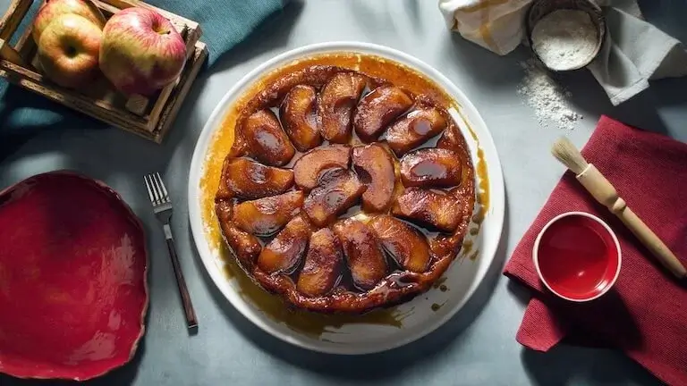 Freshly baked apple tarte Tatin with caramelized apples on a white plate beside baking tools and apples.