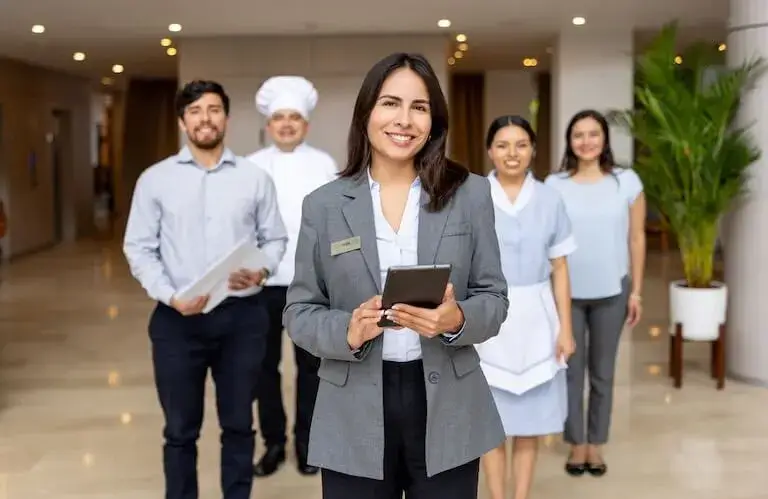 Hospitality manager in gray business suit holding tablet stands confidently in modern restaurant lobby with diverse team of hospitality staff including chef and servers in background.
