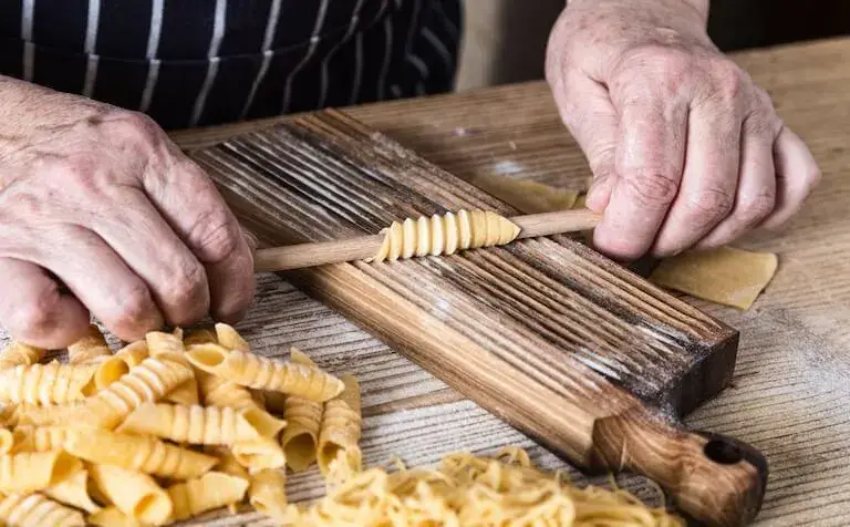 Close-up of hands shaping homemade pasta with a wooden tool on a floured surface.