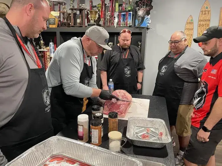 Kevin demonstrating how to trim meat in a classroom setting surrounded by students wearing aprons.
