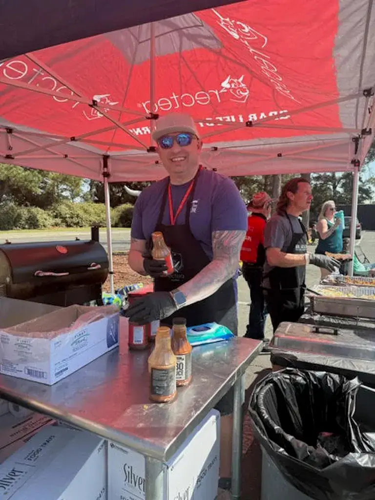 Kevin smiling under a red canopy while preparing sauces and food at a barbecue competition or event.