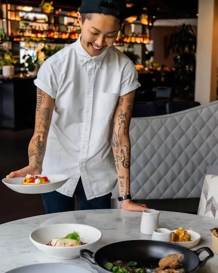 Kristen Kish stands at a table in a restaurant, smiling and holding a white dish with a meal.