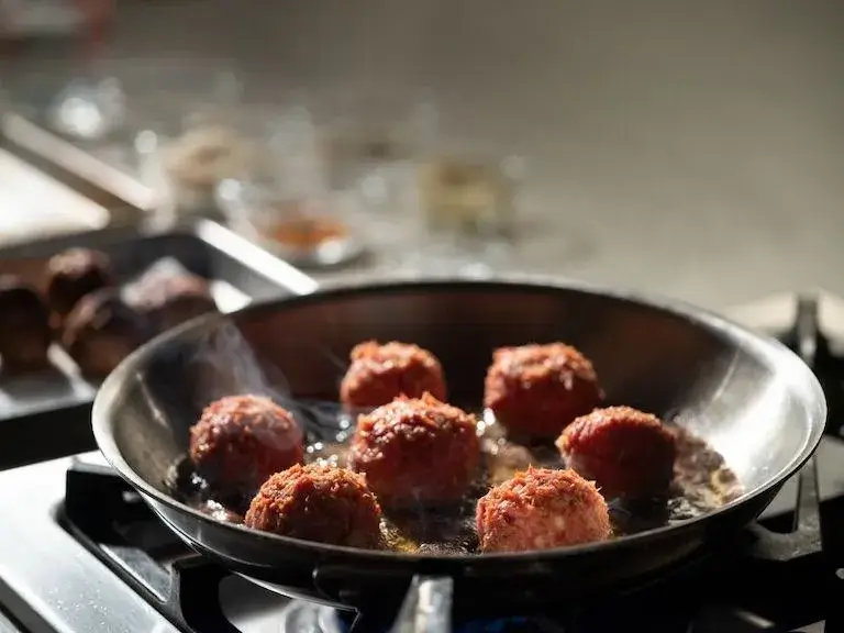 Meatballs cooking in a skillet over a gas burner with steam rising.