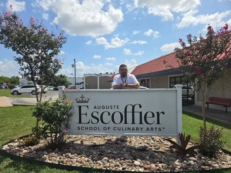 Mike Carrillo leans on a sign reading Auguste Escoffier School of Culinary Arts at the Austin Campus.