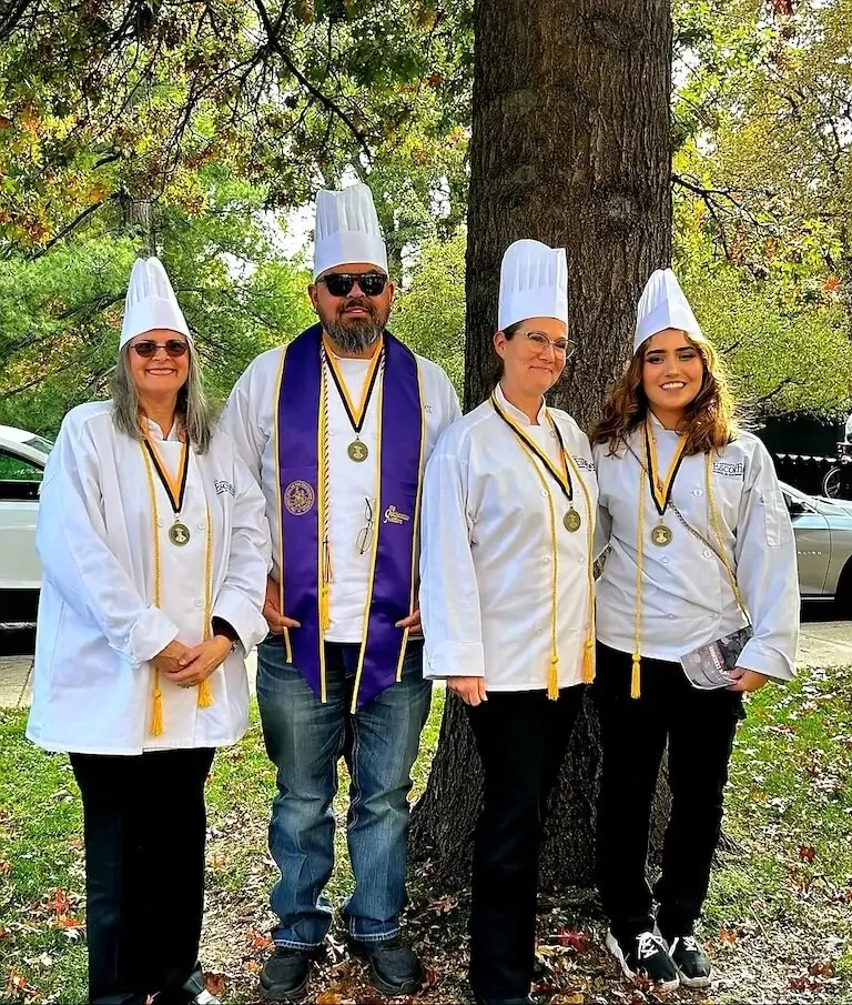 Mike Carrillo poses outdoors alongside other Escoffier graduates, all wearing chef’s coats, toques, and medallions.