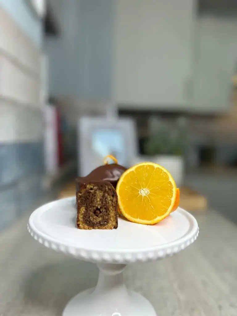 Orange swirl cake with chocolate glaze, next to a sliced orange on a white cake stand.