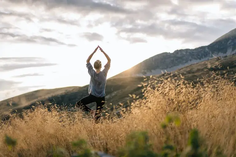 Person practicing yoga in a grassy field with mountains in the background, symbolizing wellness and balance.
