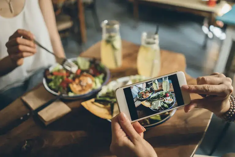Person taking a photo of a colorful salad and drinks at a restaurant table with a smartphone.