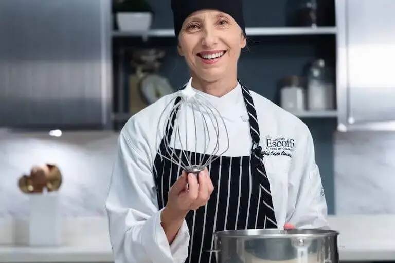 Smiling chef in Escoffier uniform holding a wire whisk in a kitchen setting.