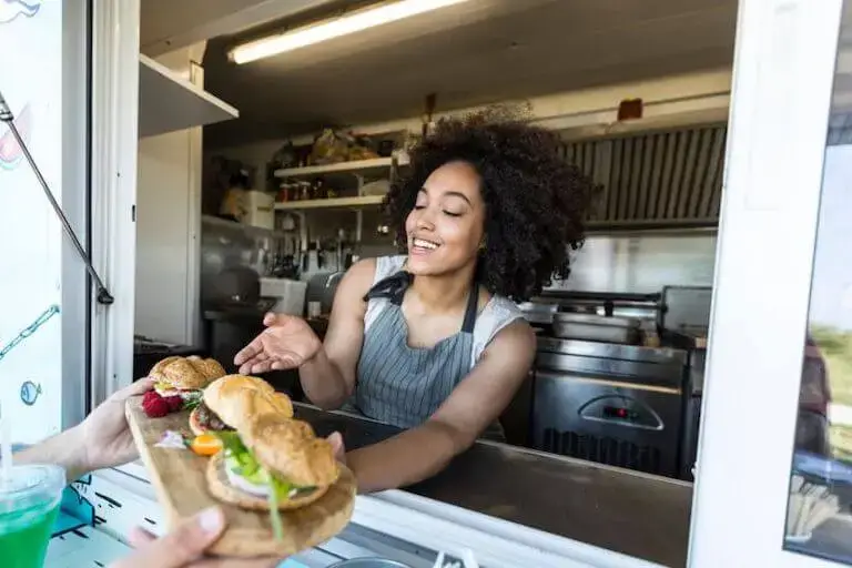 Smiling woman handing out gourmet sandwiches from a food truck window.