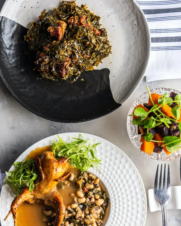 Smothered quail served on a white dish and collard greens served on a black and white dish on a white counter with napkin and fork.