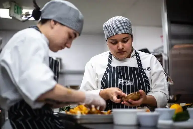 Two culinary students in uniform prepare ingredients in a professional kitchen, one focused on peeling a vegetable while the other works nearby.