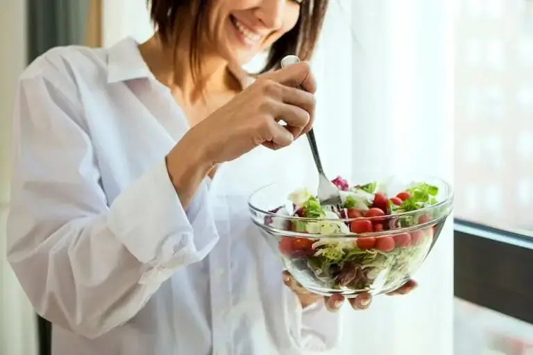 Smiling person in a white shirt holding a glass bowl of salad with lettuce and cherry tomatoes near a window.